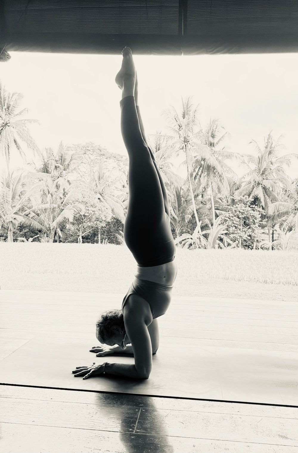 Gretchen Ramsey in a full standing handstand on an open-air deck, palm trees and rice fields behind her